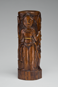 A small cylinder of shiny, dark brown wood. Carved on it is a woman styled to look like an ancient cave drawing.